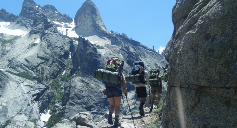 Three people wearing backpacks approach a rocky summit under blue skies